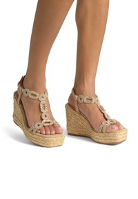 Beige Keilabsätze mit geflochtenen Jutesohlen, ausgestattet mit einem verzierte Riemendesign und einem Knöchelverschluss. Offene Zehenform mit dekorativen Akzenten.