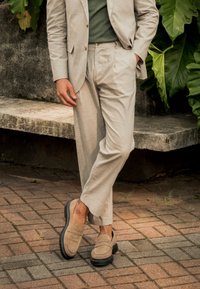 Traje de color beige claro con un diseño ajustado, combinado con una camisa verde y unos zapatos de lona beige. El entorno incluye una superficie de piedra texturizada y vegetación.