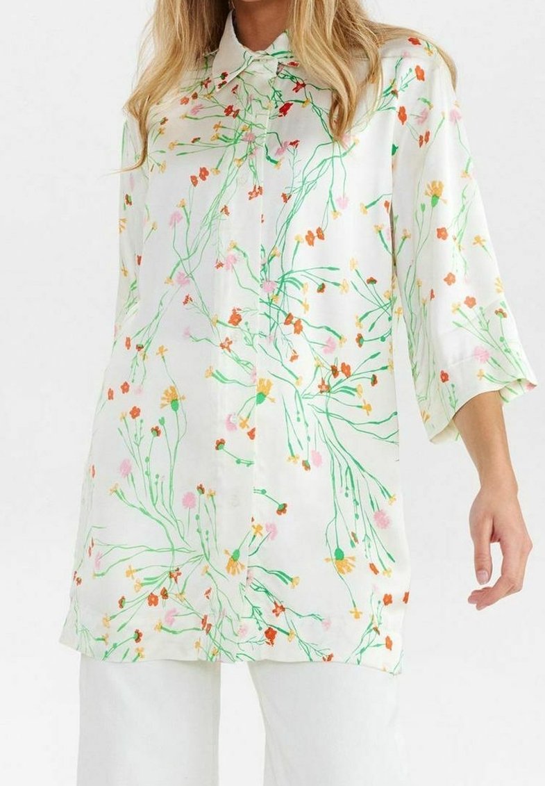 Blouse blanche à manches longues avec imprimé floral multicolore, fermeture à boutons à l'avant, portée avec un pantalon blanc.