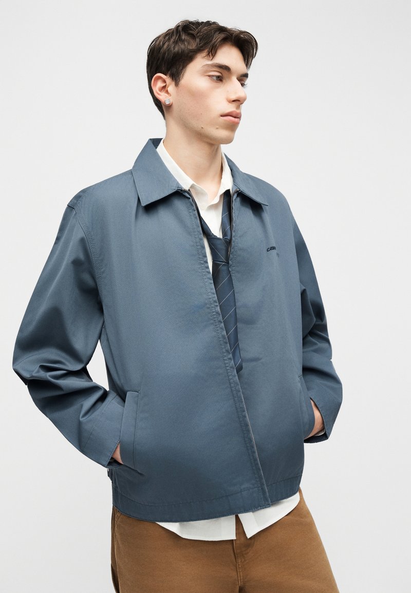 Giovane uomo con capelli corti e scuri che indossa una giacca blu con zip, camicia bianca, cravatta blu e pantaloni marroni, con le mani nelle tasche della giacca, che guarda di lato.