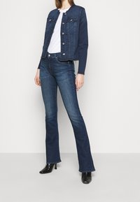 Chaqueta de mezclilla oscura con botones plateados, dos bolsillos en el pecho y dobladillo deshilachado. Combinada con jeans oscuros acampanados y botas de tacón negras.