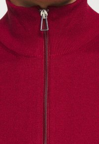 Punainen ribbipintainen turtleneck-neule, jossa on puolivetoketju, sileä rakenne ja metalliset vetoketjivarusteet, suunniteltu istuvaksi.