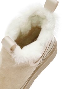 Stivale alla caviglia in suede beige con una fodera in pelliccia sintetica bianca. Presenta una superficie morbida e una linguetta in tessuto a forma di anello per una facile calzata. Suola a profilo basso.