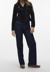 Svart mockalädermotorcykeljacka med silverdetaljer och dragkedjor, tillsammans med högmidjade mörka denimbyxor med vida ben och bruna sneakers.