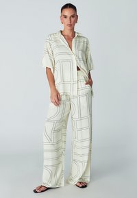 Vrouw in crèmekleurige, losse broek en bijpassende knoopblouse met zwarte geometrische lijnpatronen, gecombineerd met zwarte sandalen.