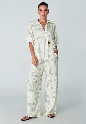 Vrouw in crèmekleurige, losse broek en bijpassende knoopblouse met zwarte geometrische lijnpatronen, gecombineerd met zwarte sandalen.