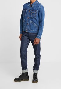 Modrá džínová bunda s náprsními kapsami, hnědými knoflíky a kontrastním šitím; v kombinaci s tmavými džínami a černými kotníkovými botami.