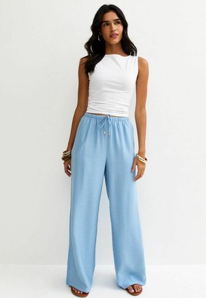 Młoda kobieta o ciemnych włosach ubrana w białą, bezrękawnikową bluzkę, jasnoniebieskie szerokie spodnie z troczkiem, bransoletki i sandały, stoi na tle białej ściany.