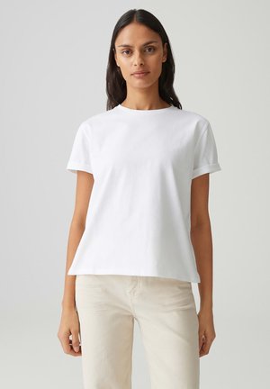 Frau mit langen dunklen Haaren, die ein schlichtes weißes T-Shirt mit kurzen Ärmeln und hellbeige Hosen trägt, steht vor einem neutralen Hintergrund.