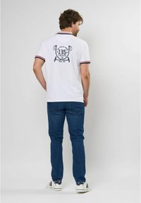 Hvid poloshirt med et sort grafisk print på ryggen, prydet med krave og ærme accenter i rød og marineblå striber. Bæres med blå jeans.