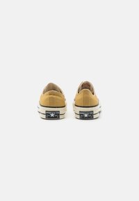 Gul lærred lave sneakers med en hvid gummisål og sort hælpatch, der har "Converse All Star" logoet.