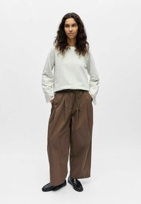 Femme aux cheveux bouclés portant un haut à manches longues gris clair avec des détails en dentelle, un pantalon large marron et des mocassins noirs, debout devant un fond uni.