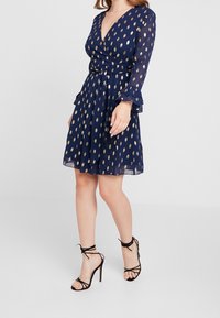 Sjöblå omslagklänning med guldprickigt mönster, långa ärmar, volangärmar och figursydd midja. Matchas med svarta sandaler med hög klack och remmar.