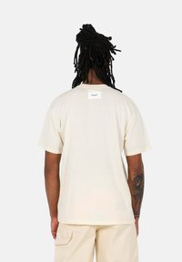 Crème-kleurig t-shirt met korte mouwen en een geweven label op de achterzijde, getoond vanaf de achterkant, met een ontspannen pasvorm en een zachte textuur.