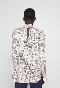 Persona che indossa una blusa bianca a maniche lunghe con pois neri, dettaglio a chiave sul retro e due bottoni per fissarla al collo.