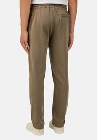 Chaki jogger kelnės su elastinga juosmena, glotnia tekstūra, viena užpakalinė kišenė ir siaurėjantis kojų dizainas, dėvimos su béžiniais batais.