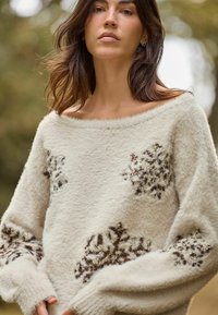 Cremefarbener, flauschiger Pullover mit schulterfreiem Design. Merkmale sind braune und silberne Schneeflockenmuster. Lange Ärmel mit gerippten Bündchen.