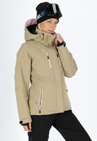 Giacca impermeabile beige con dettagli in zip rosa, cappuccio aderente e polsini neri. Indossata con pantaloni neri e un berretto nero.