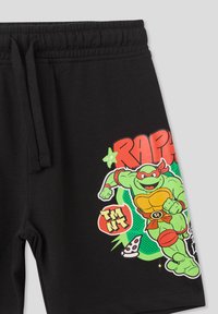 Shorts di cotone neri con una grafica vivace di un personaggio tartaruga dai toni verdi con accenti rossi, testo "RAPHAEL" e dettagli a forma di fetta di pizza.