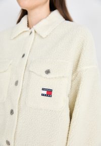 Kreem värvi fliisjakki, millel on kraega disain, kahte esist taskut ja "TOMMY JEANS" logoga riidepatch. Ees on hõbedased nupud.
