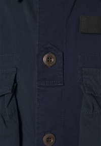 Merensininen paita, jossa on napillinen edusta, kaksi päällystaskua, kuusikulmainen nappidetalji ja näkyvät saumat teksturoidussa kankaassa.