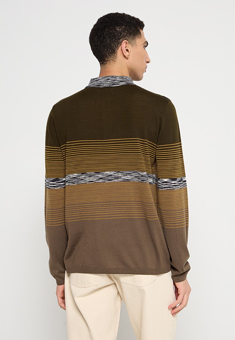 Mann, der von hinten steht und einen gestreiften braun-gelben Pullover mit beigen Hosen trägt, vor einem schlichten Hintergrund.