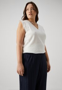 White sleeveless top with a textured fabric and scalloped lace trim, paired with dark blue pleated wide-leg pants.