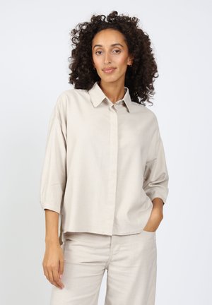 Beige oversized shirt with a pointed collar, button closure, and three-quarter sleeves. Smooth texture, casual design, worn with matching pants.