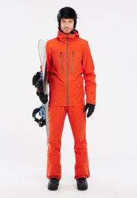 Giacca e pantaloni da snowboard di un vivace arancione, dotati di tasche con zip e guanti. Snowboard nero con attacchi blu sostenuto su uno sfondo neutro.