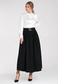 Weißes Satin-Blusenhemd mit Kragen, kombiniert mit einem schwarzen, plissierten Maxirock. Das Outfit wird von einem schwarzen Gürtel mit runden silbernen Akzenten und hohen Absätzen ergänzt.