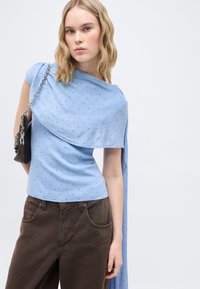 Ξανθιά γυναίκα που φοράει ένα ανοιχτό μπλε, ασυμμετρικό μπλουζάκι με πουά σχέδιο και μακριά πτυχή, σκούρο καφέ παντελόνι, και κρατάει μια μαύρη τσάντα με λουρί αλυσίδα στον ώμο.