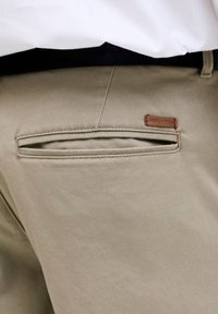 Nærbilde av baklommen på beige bukser med sort belte og hvit skjorte stukket inn, med en liten brun "Jack & Jones"-etikett.