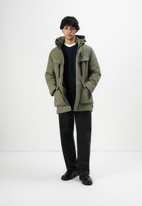 Junger Mann, der eine olivgrüne Kapuzenjacke, schwarze Hosen und schwarze Stiefel trägt, mit Händen in den Taschen, vor einem schlichten weißen Hintergrund steht.