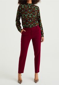 Långärmad blus med hög hals i gröna och bruna abstrakta mönster, tillsammans med smala burgundy-byxor och beige spetsiga klackar.