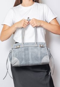 Borsa in suede grigia con accenti in pelle, manici doppi e nappine laterali. Dettagli notevoli includono una fibbia e bordi arrotondati.