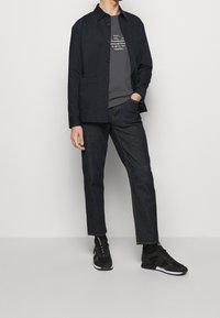 Marinblå knäppt jacka över en mörkgrå grafisk t-shirt, tillsammans med mörk denimjeans och svarta sneakers med vita detaljer.