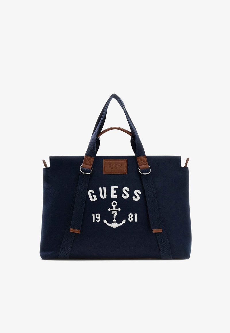 Námornícka modrá taška Guess na rameno s dvoma rukoväťami, hnedé kožené akcenty, biely kotvičkový logo a text "Guess 1981" na prednej strane.