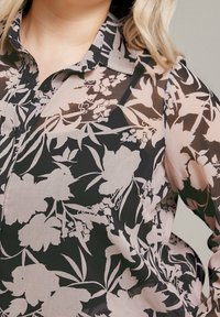 Genomskinlig blommig blus med svart bakgrund och rosa samt vita blommönster, krage och knäppningar.