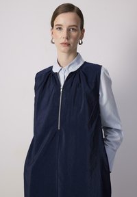 Vestido sin mangas de color azul marino con un acabado texturizado, cierre de cremallera en la parte delantera, escote con fruncido y bolsillos laterales. Sobre una camiseta de color azul claro.