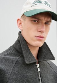 Giacca di lana grigio scuro con un colletto alto e ripiegato e una zip argentata. Un berretto beige con dettagli verdi riporta la scritta "First Class", indossato in posizione frontale.