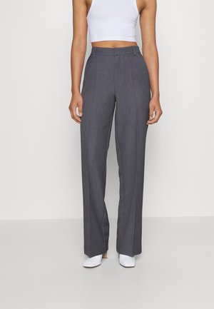 Kvinde iført højtaljede, vidde grå bukser kombineret med en hvid cropped tanktop og hvide hælede sko, stående mod en ensfarvet baggrund.