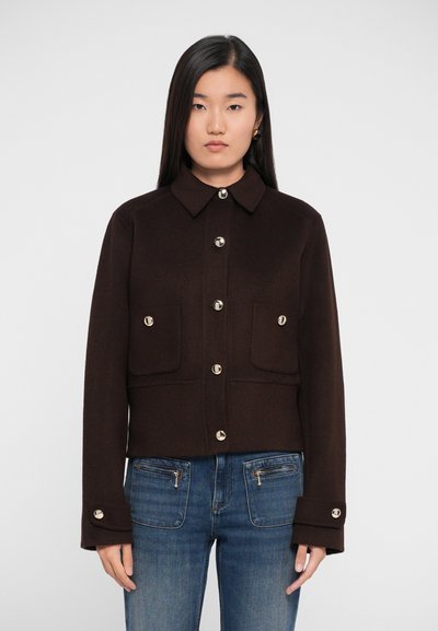 Young woman with straight black hair wears dark brown buttoned jacket with front pockets and blue jeans with zippered front pockets.