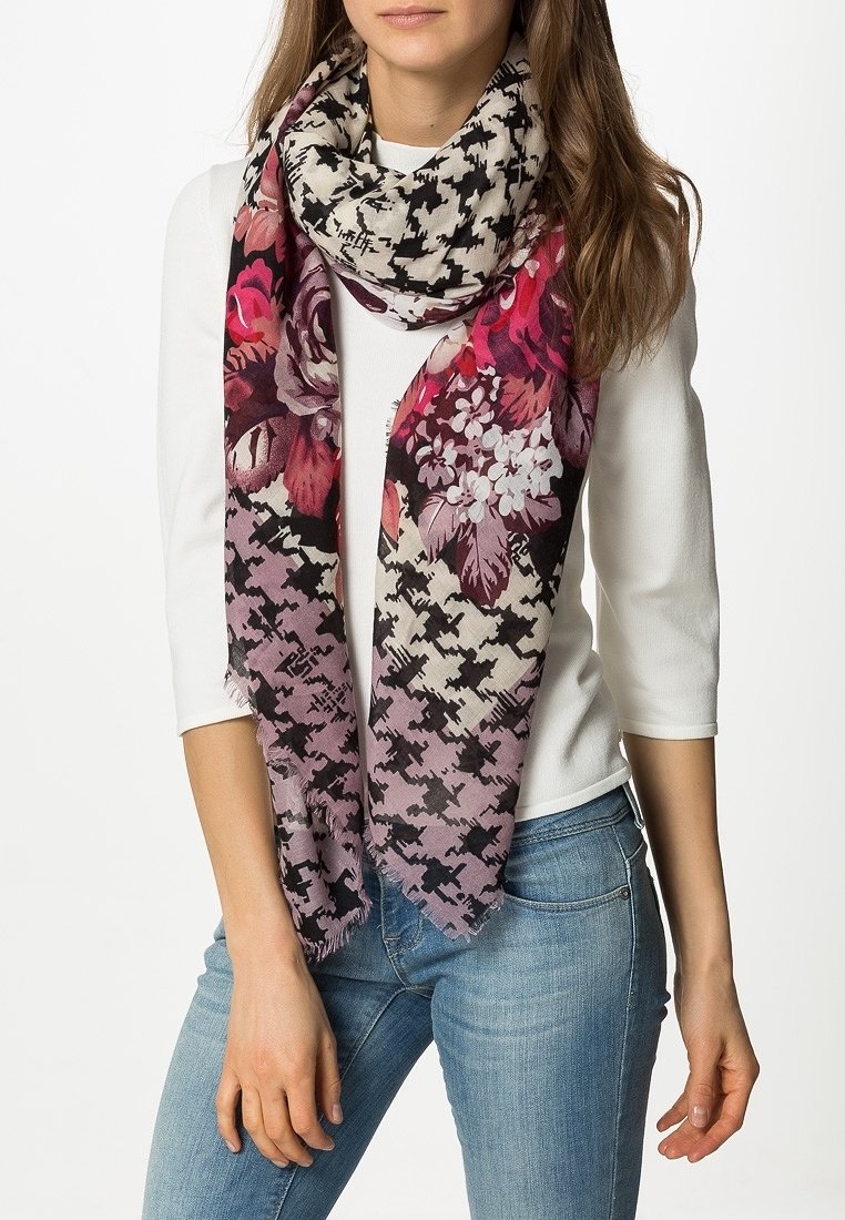 Femme portant un haut blanc, un jean bleu et une grande écharpe avec des motifs floraux et pied-de-poule rose, violet et noir enroulée autour du cou.