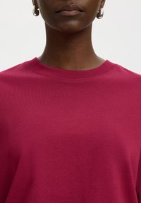 Κοντινό πλάνο ενός ατόμου που φοράει μονόχρωμο σκούρο ροζ μπλουζάκι με στρογγυλή λαιμόκοψη και ασημένια κρίκους σκουλαρίκια σε λευκό φόντο.