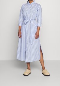 Gestreepte button-up jurk in blauw en wit met een strakke taille, 3/4 mouwen en zijslitsen, gecombineerd met beige instapschoenen.