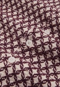 Tessuto crema con motivo geometrico testurizzato ricamato in viola scuro e tre bottoni bianchi cuciti sopra.