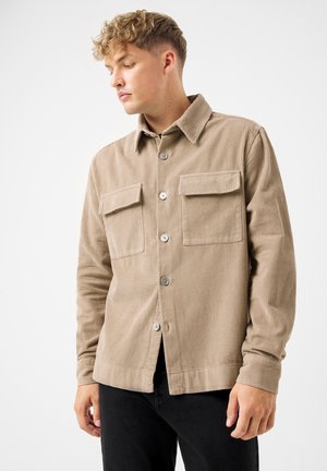 Camisa de pana beige con cuello, dos bolsillos en el pecho y botones plateados. Presenta una textura acanalada y un ajuste holgado. Pantalones negros visibles.