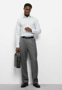 Hellblau kariertes Hemd, graue Hose, schwarzer Gürtel und schwarze Lederschuhe. Hält eine schwarze Aktentasche und steht vor einem weißen Hintergrund.