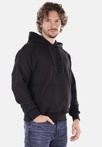 Felpa nera in cotone con tasca a marsupio, cappuccio con stringa e polsini e orlo a costine; il modello indossa jeans blu.