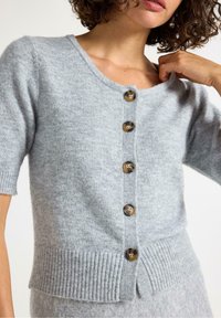 En kvinde med krøllet hår justerer en lysegrå cardigan med fem brune knapper og en ribbet kant, der fremhæver en afslappet fashionstil.